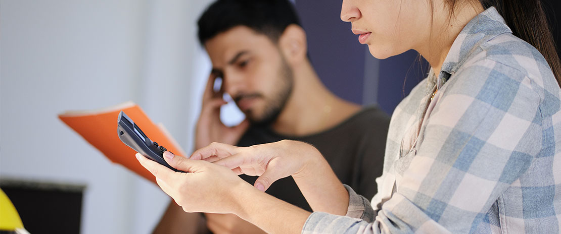 Mulher observa um telefone fixo sem fio. Atrás, desfocado há um homem pensativo.