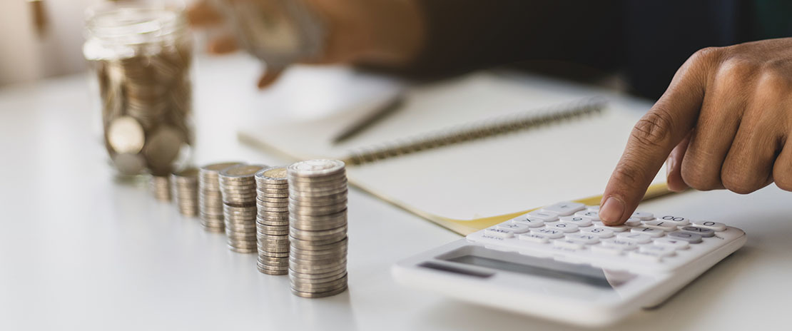 Diversas pilhas de moedas e uma mão tocando uma calculadora branca.