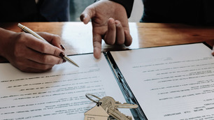 Mão indica para outra mão onde assinar em um contrato. Acima do papel desse contrato há um molho de chaves.