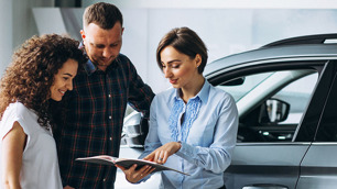 Mulher mostrando a um casal algo em uma revista. Atrás há um carro.