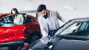 Homem entrando em um carro em uma concessionária. Ao fundo uma mulher faz o mesmo em outro veículo.