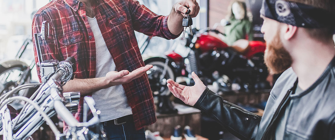 Homem entrega uma chave de moto para outro homem em meio a outras motos.