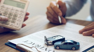 Uma mão com uma caneta se aproxima de uma folha, enquanto em cima dessa folha há uma controle de carro e um carro em miniatura.