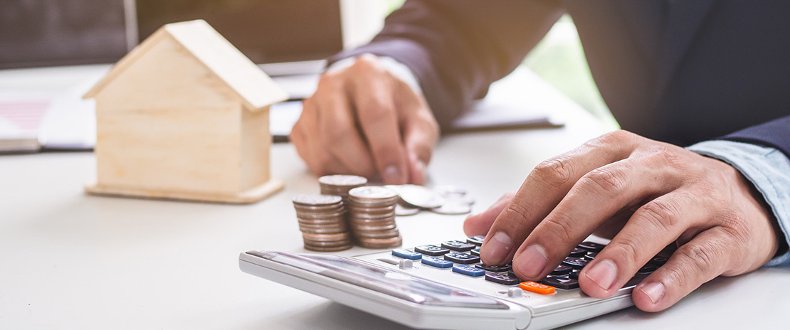 Mãos masculinas teclam uma calculadora. Há também uma pilha de moedas e uma casa em miniatura.