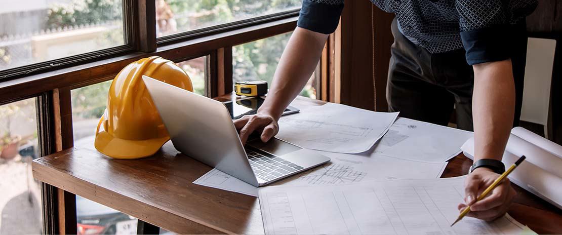 Homem observa um notebook e diversas folhas em uma mesa. Ao lado ainda há um capacete de obra.