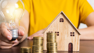 Homem vestido de camiseta amarela empunha uma lâmpada em uma mão e á frente dele há uma casa em miniatura e pilhas de moedas.